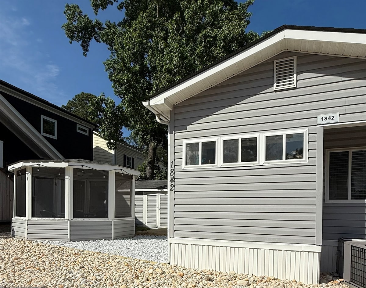An exterior view of a charming bungalow features light gray siding and multiple windows. A spacious gazebo with screens is positioned next to the home, providing a shaded outdoor seating area. Gravel landscaping complements the structure against a blue sky.