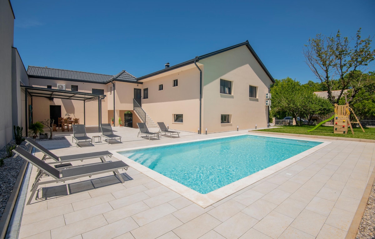 A spacious outdoor area features a private pool surrounded by sun loungers. The modern holiday home stands in the background, with large windows and a cozy dining space visible. A playground area is included, set against a backdrop of green trees.