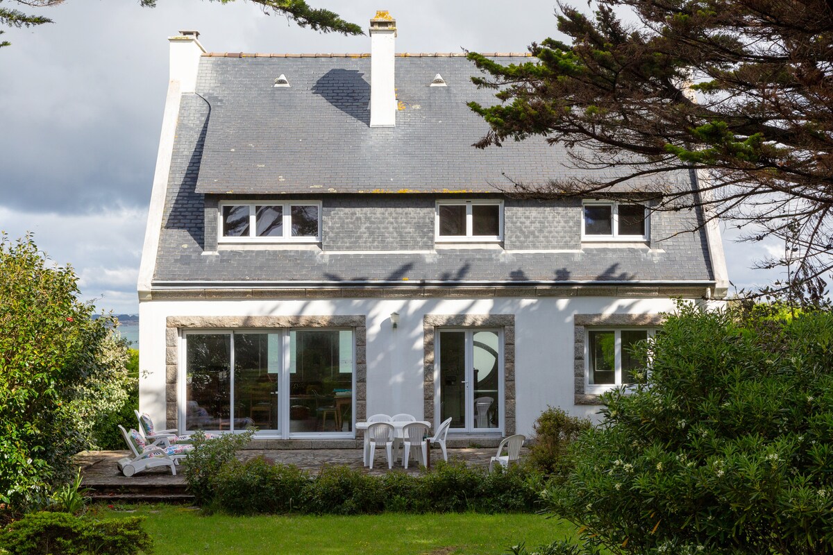 The exterior of the holiday home presents a two-story structure with a combination of slate and white facade. Large windows provide ample natural light to the interior. A small patio area with several white chairs is visible, surrounded by lush greenery and trees.