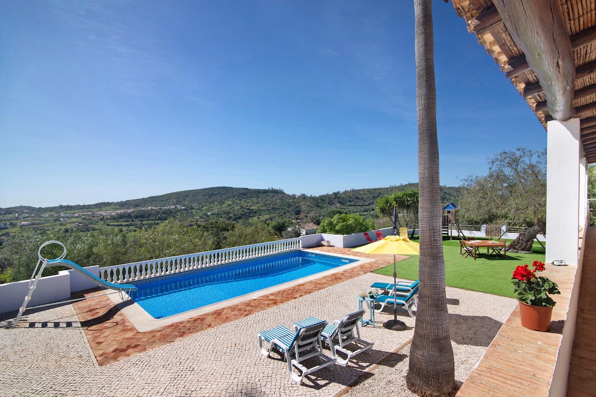 An outdoor area is highlighted by a private swimming pool surrounded by lounge chairs. A grassy section features a playground and a shaded seating area. Lush hills are visible in the background, under a clear blue sky.