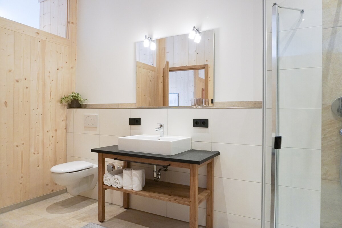 A modern bathroom is featured, highlighting a stylish sink atop a wooden vanity with rolled towels. Natural light enters through a high window, and sleek mirrors enhance the clean aesthetic. A glass shower enclosure and a toilet complete the space.