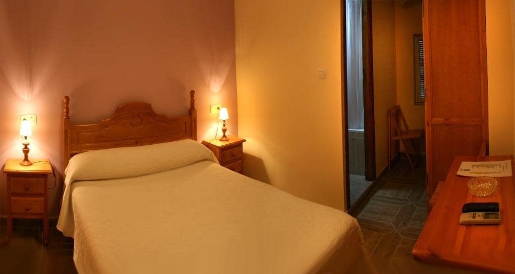 A cozy sleeping area features a wooden bed with a light-colored bedspread. Two bedside tables with lamps provide soft lighting. The wall is painted in a warm hue, and a small wooden desk is visible in the room. A doorway leads to a bathroom.