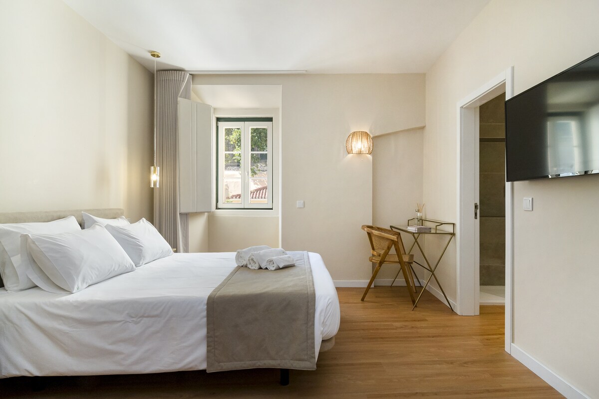 The cozy sleeping area features a spacious bed with soft linens and decorative pillows. A work desk and chair are positioned nearby, complemented by a stylish lamp. Natural light enters through a window with an unobstructed view. The modern bathroom is accessible through a door.