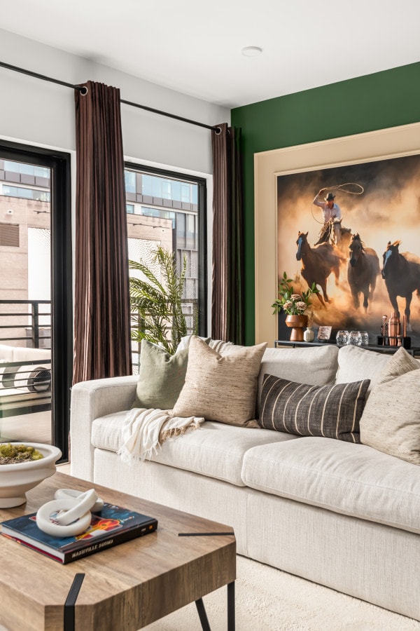 The living area presents a neutral-colored sofa adorned with decorative pillows, accompanied by a coffee table. Two large windows with dark curtains allow natural light to enter while providing views of the outdoor balcony. In the background, a striking western-themed artwork features galloping horses.