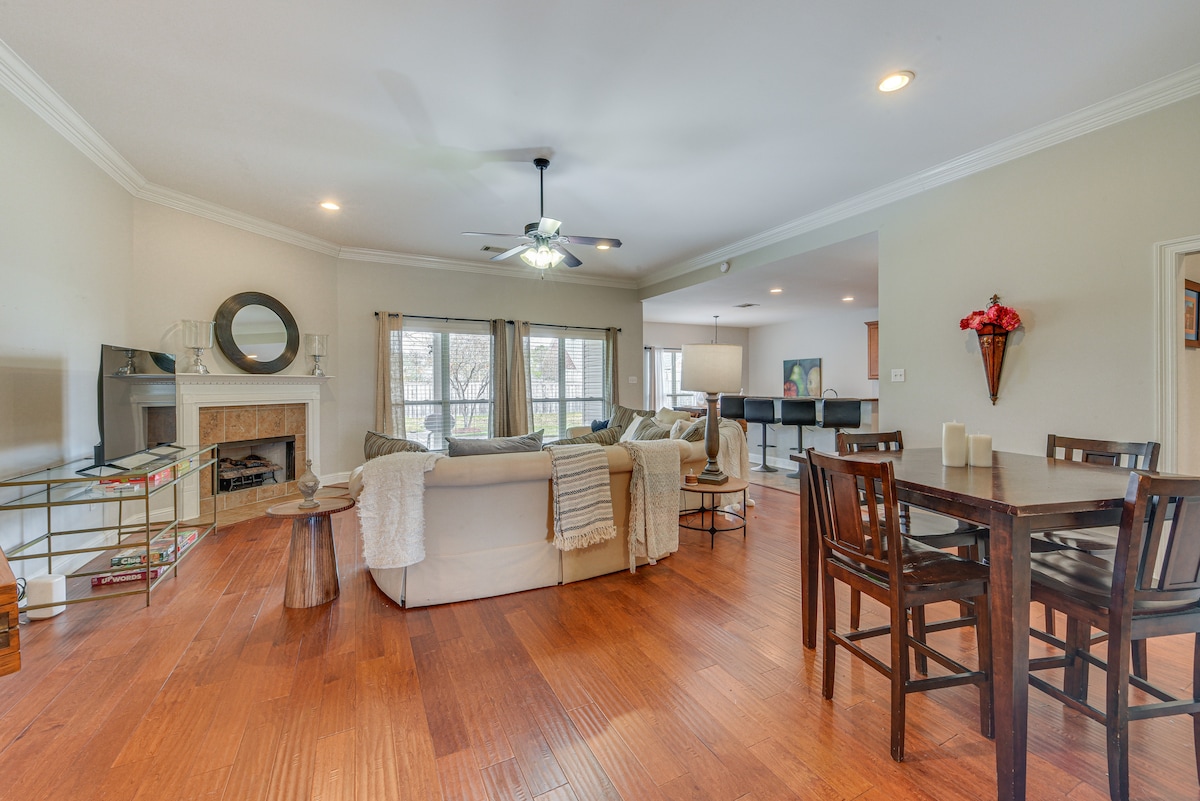 An open living area features wooden flooring, a ceiling fan, and natural light from large windows. A comfortable sofa adorned with throw pillows is positioned near a fireplace, while a dining table for four stands nearby, contributing to a welcoming atmosphere.