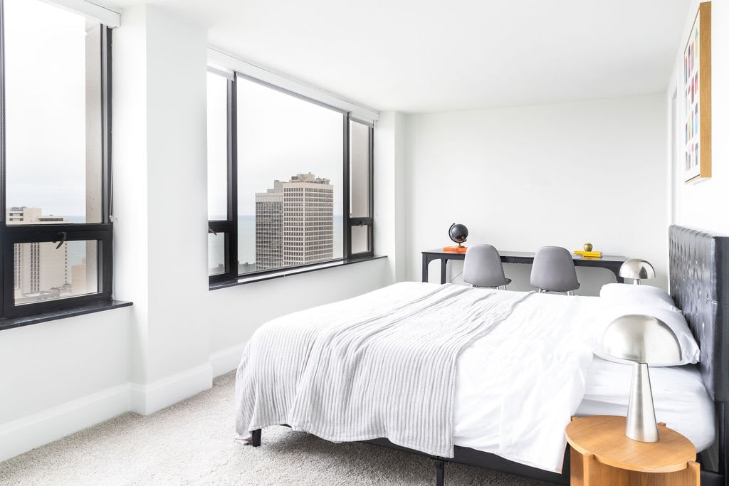 A well-lit bedroom features large windows offering expansive views of the cityscape. A spacious bed is dressed in white linens, accompanied by a light grey throw. A small desk with two chairs sits nearby, complemented by modern table lamps.