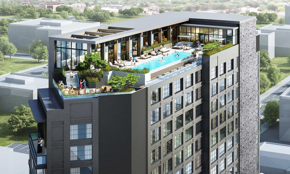 A modern high-rise building showcases a rooftop outdoor pool area surrounded by lounge chairs and greenery. Large glass windows provide views of the surrounding landscape, emphasizing the building's contemporary design. The rooftop space is set up for relaxation and socializing.
