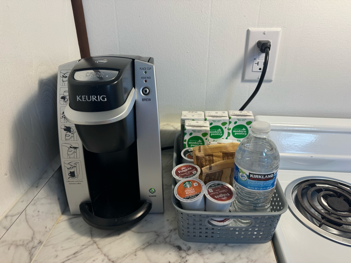 A Keurig coffee maker is displayed on a marble countertop, accompanied by a selection of coffee pods in various flavors. A basket contains creamers and sweeteners, with a bottle of water placed nearby. The kitchen appliance setup offers convenience for guests.