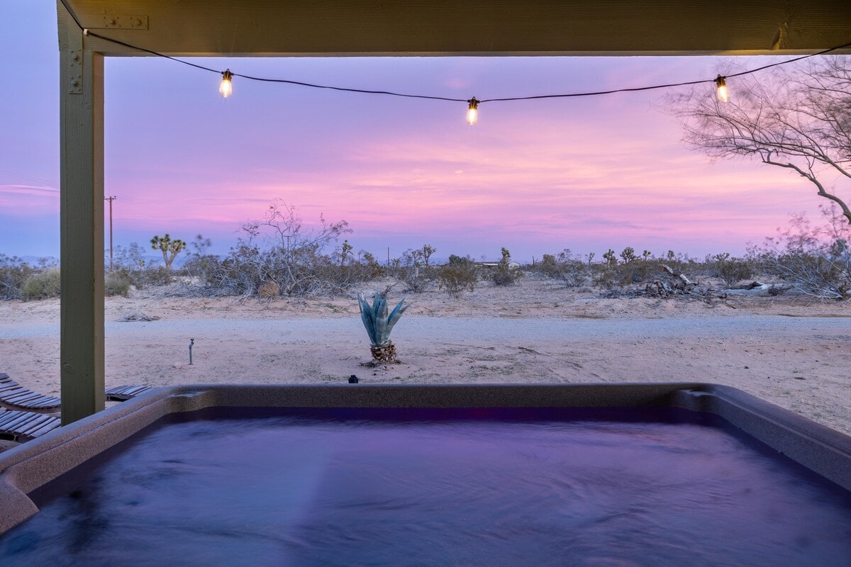 A hot tub is situated beneath a covered patio, providing a serene view of the desert landscape. The sunset paints the sky in soft purple and pink hues, while warm lights create a cozy ambiance against the backdrop of Joshua trees and arid terrain.