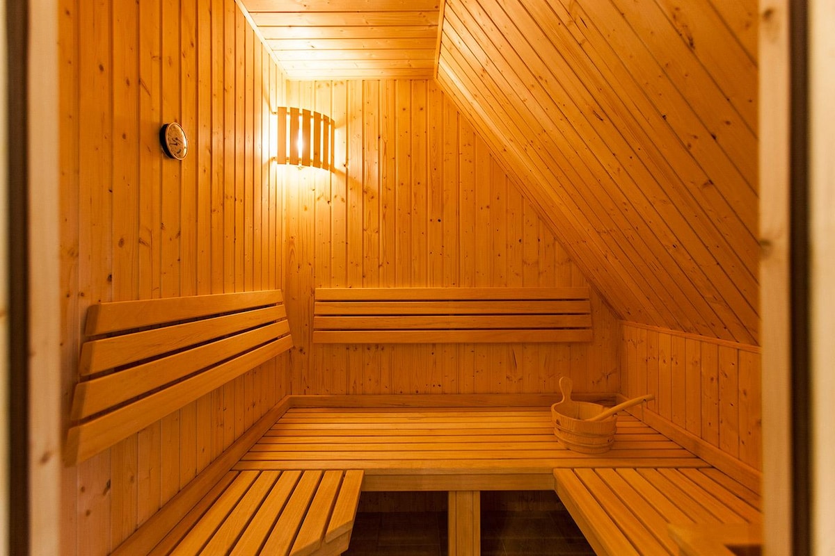 The sauna features wooden paneling and benches, creating a warm and inviting space. Soft lighting is provided by wall-mounted fixtures, enhancing the relaxing atmosphere. A wooden bucket is visible, ready for use, while a thermostat control is mounted on the wall.