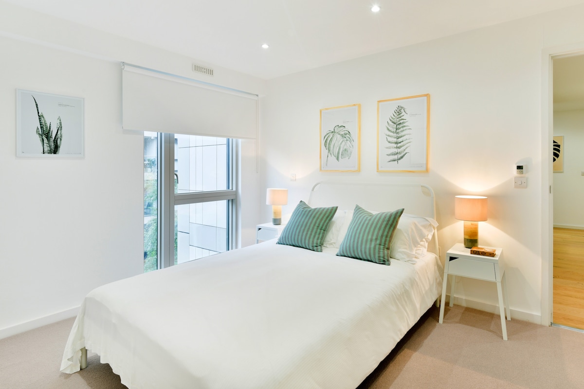 A bright bedroom is presented with a spacious bed adorned with striped cushions. Two bedside tables hold softly glowing lamps, providing gentle lighting. Botanical artwork hangs on the pale walls, and a large window allows natural light to fill the room.