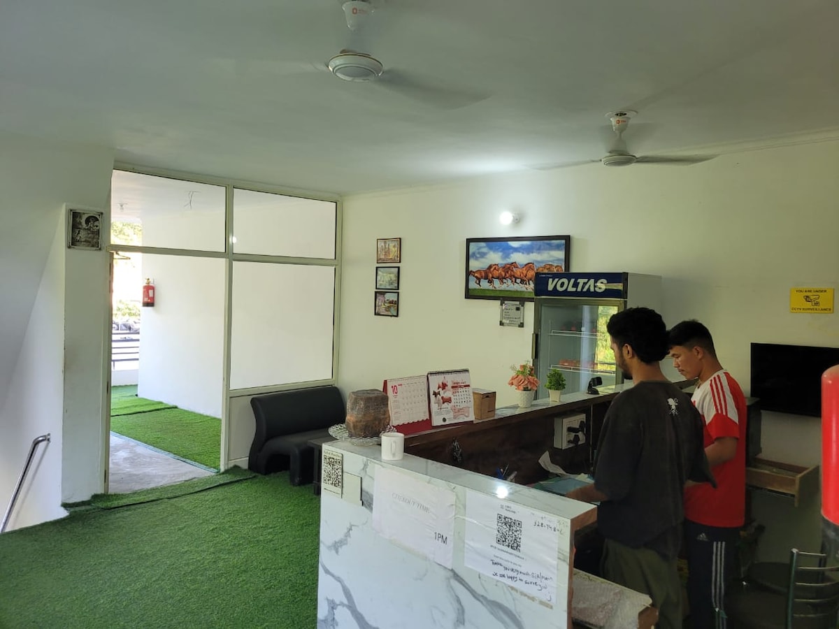 A reception area features a marble-topped counter with two individuals attending to guests. The space is decorated with framed artwork and a plant, while a door leads to a green carpeted entryway. Light from large windows brightens the room.
