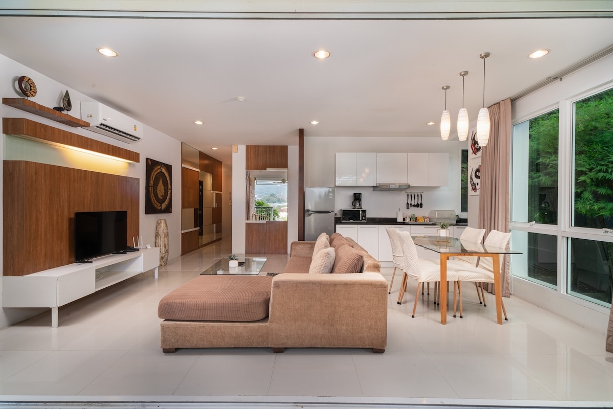 A modern living area features a spacious sectional sofa, a sleek television console, and a dining table with white chairs. Large glass doors allow natural light to fill the room, offering views of the lush surroundings. The kitchen is equipped with contemporary cabinetry and appliances.
