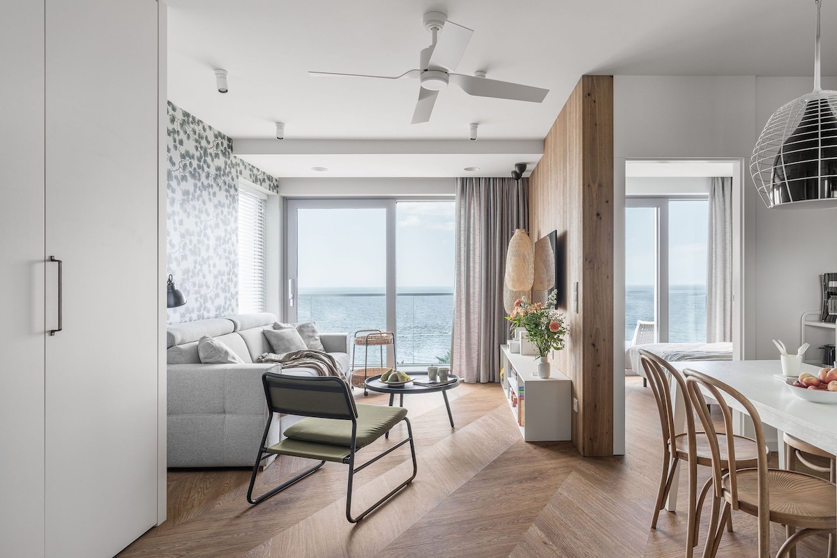A bright open-plan living area features large sliding glass doors providing a view of the sea. A comfortable gray sofa and stylish chairs surround a coffee table. The modern kitchenette is visible, complemented by natural wood accents and light flooring.