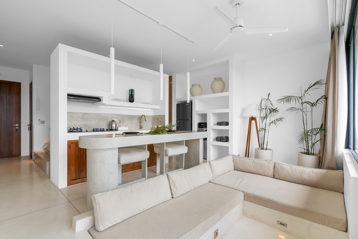 An open-plan living area features a sunken lounge with a soft neutral-toned sofa. A modern kitchen is equipped with premium appliances and a sleek countertop. Large windows allow natural light to brighten the space, complemented by minimalist decor and indoor greenery.