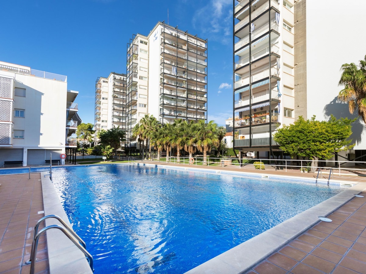 A large swimming pool is shown, surrounded by palm trees and landscaped green areas. The clear blue water reflects sunlight, and there are buildings in the background. The pool area includes paved decking and steps leading into the water.