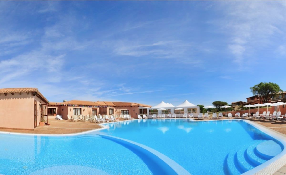 The image presents a panoramic view of a spacious swimming pool surrounded by lounge chairs and tents. The pool features an inviting blue hue under a clear sky, with nearby buildings reflecting a traditional architectural style, set within a tranquil outdoor environment.