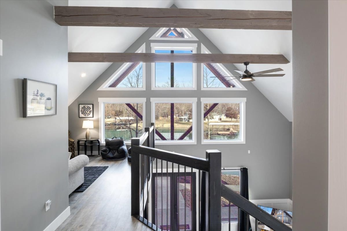 The upstairs living area features a spacious design highlighted by architectural beams and large triangular windows. Natural light fills the room, offering views of the surrounding landscape. A cozy seating area includes a comfortable chair and a sofa, creating an inviting space for relaxation.
