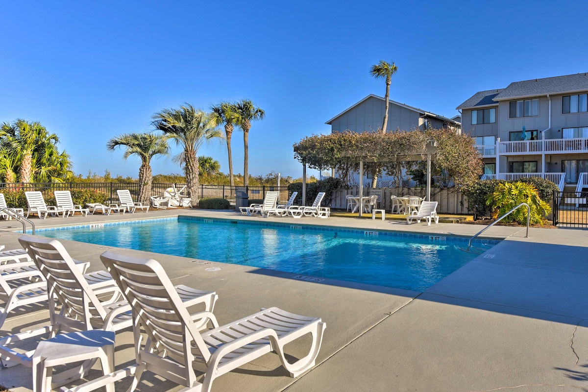 A spacious pool area is surrounded by lounge chairs and tropical palm trees. The pool is inviting and well-maintained, with a clear blue surface reflecting the sunny sky. A building with balconies is visible in the background, enhancing the outdoor relaxation space.
