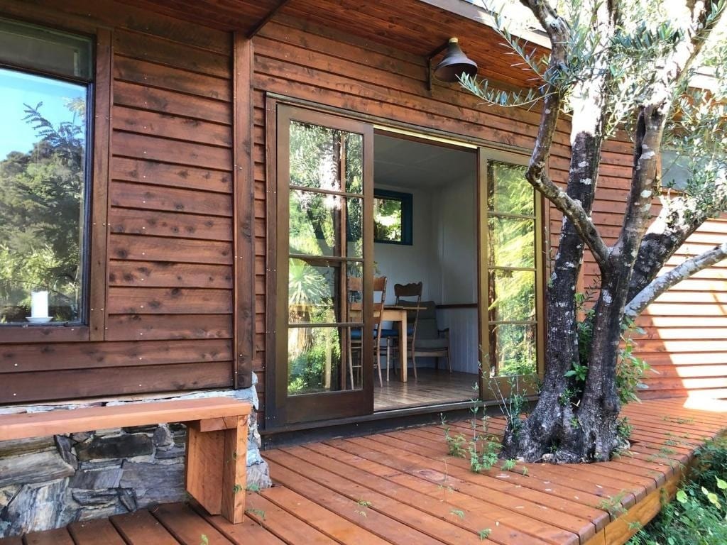 An inviting entrance features large glass doors opening onto a wooden deck. Surrounding greenery provides a natural backdrop. A small bench and low stone wall complement the exterior, while a glimpse of dining chairs is visible through the door.
