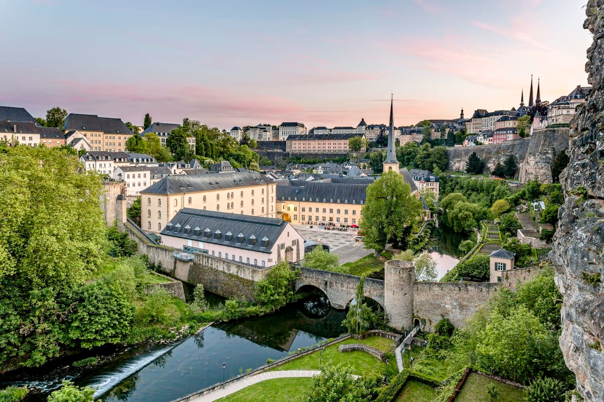 A panoramic view of a tranquil landscape features historical buildings, lush greenery, and a river. A stone bridge spans the water, connecting the banks, while gently sloping hills rise in the background, dotted with residential structures. The sky displays soft pastel colors at dusk.