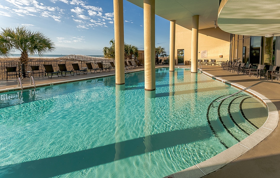 An outdoor pool area is highlighted, featuring crystal-clear water and sunlit reflections. Lounge chairs, arranged along the poolside, provide ample seating for relaxation. Palm trees sway gently in the background, enhancing the tranquil coastal setting.