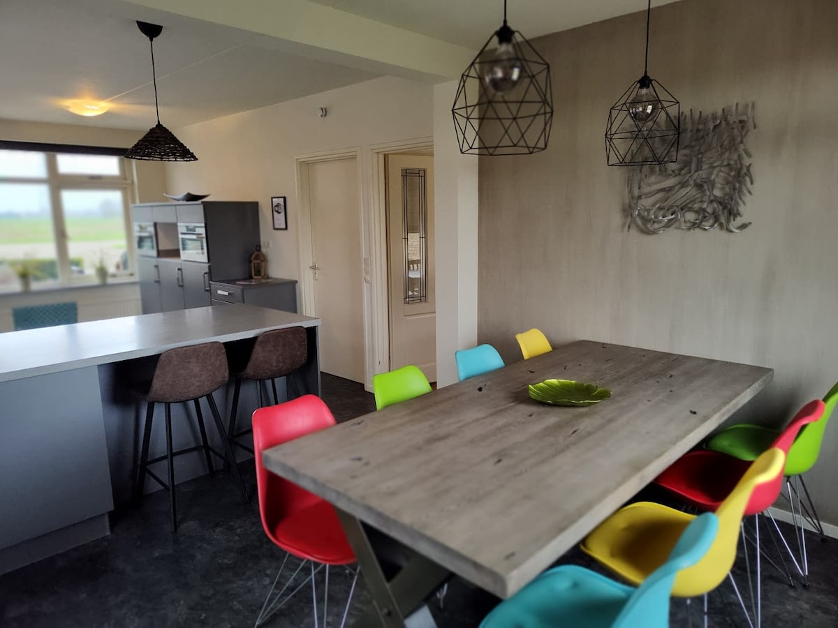 A modern kitchen and dining area features a large wooden table surrounded by colorful chairs. Unique pendant lights hang over the table, while an island bar with seating is situated nearby. Large windows allow natural light to illuminate the space.