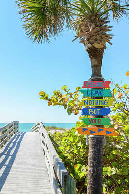 A wooden boardwalk leads toward the beach, surrounded by lush greenery and palm trees. A colorful sign is mounted on a palm tree, displaying the message 'Please Leave Nothing But Your' against a bright blue sky and the shimmering ocean in the background.