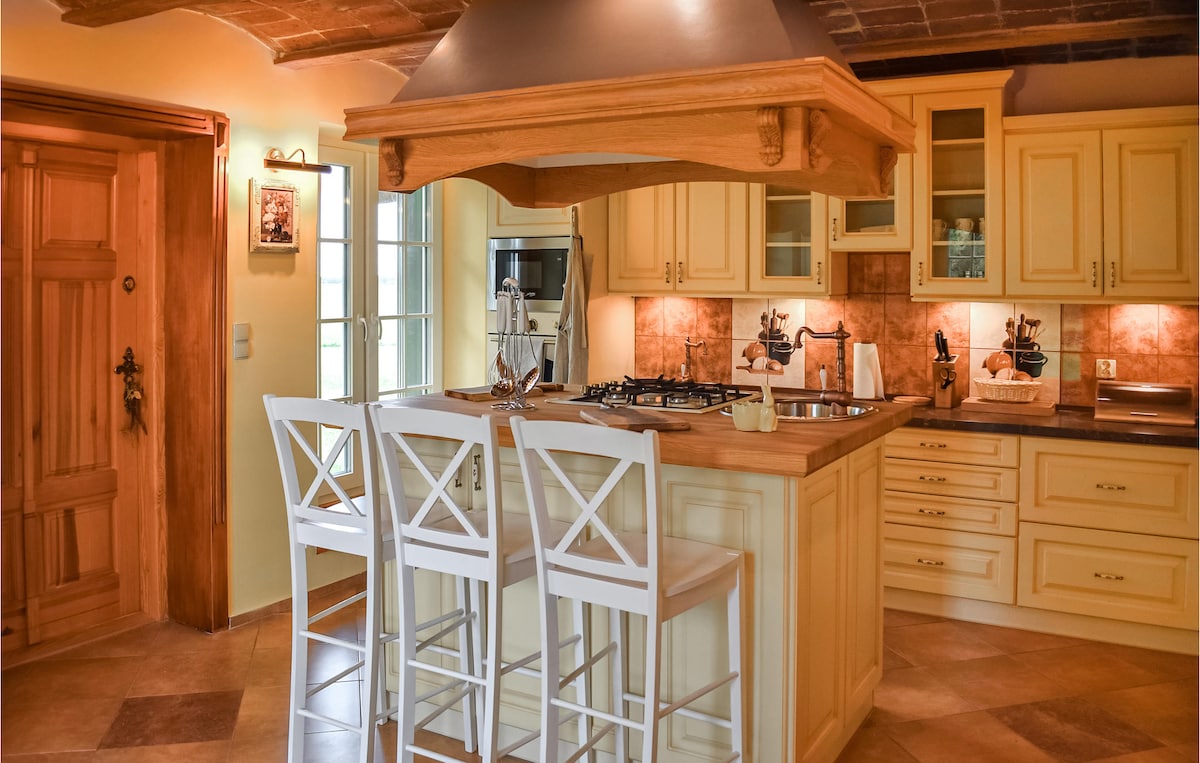 The kitchen features a central wooden island with a built-in gas stove. Two white bar stools are positioned around the island, and cabinetry in warm tones surrounds the cooking area. Natural light enters through multiple windows, illuminating the tiled floor.