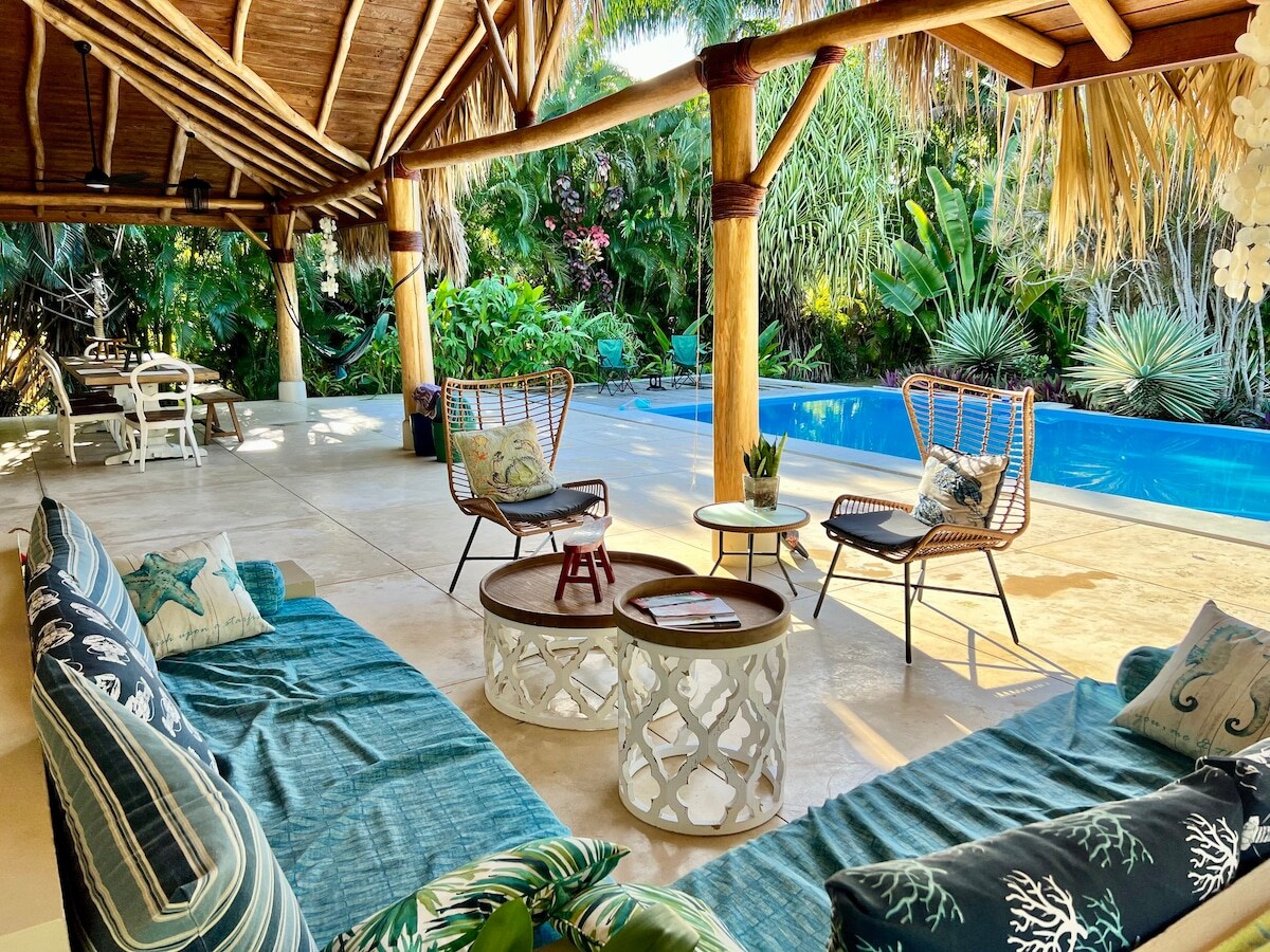 An open, covered outdoor seating area features a mix of comfortable sofas and chairs arranged around wooden tables. A private pool is framed by tropical plants and trees, offering a serene backdrop for relaxation. Natural materials are evident in the roof structure.