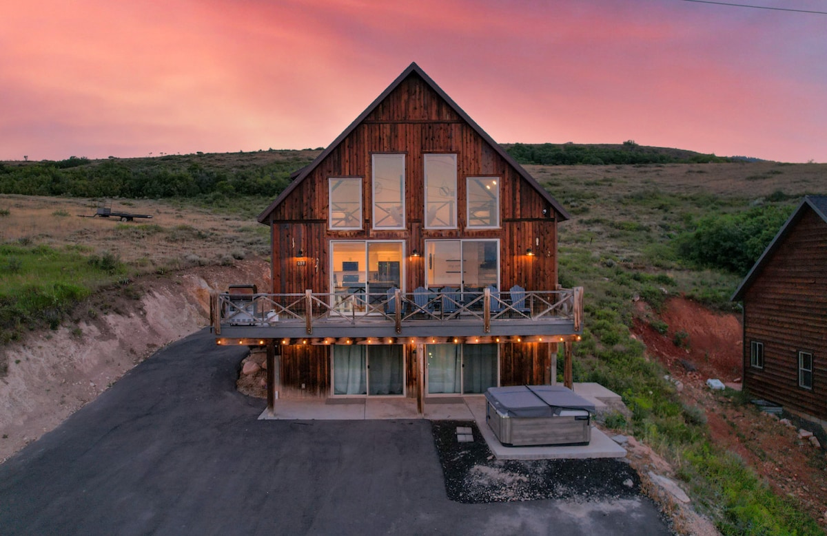 A rustic cabin is showcased against a vibrant sunset, featuring large front windows and a spacious deck with outdoor seating. A private hot tub is positioned on the patio, surrounded by grass and natural landscaping, while the paved driveway provides access to the entrance.