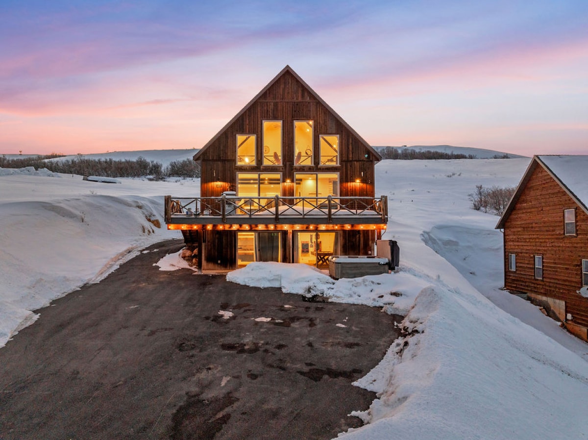 The cabin's exterior is showcased against a pastel sunset, highlighting its rustic design. Large windows provide views of the snowy landscape, while a spacious deck invites outdoor enjoyment. Paved parking area is visible, accommodating several vehicles.
