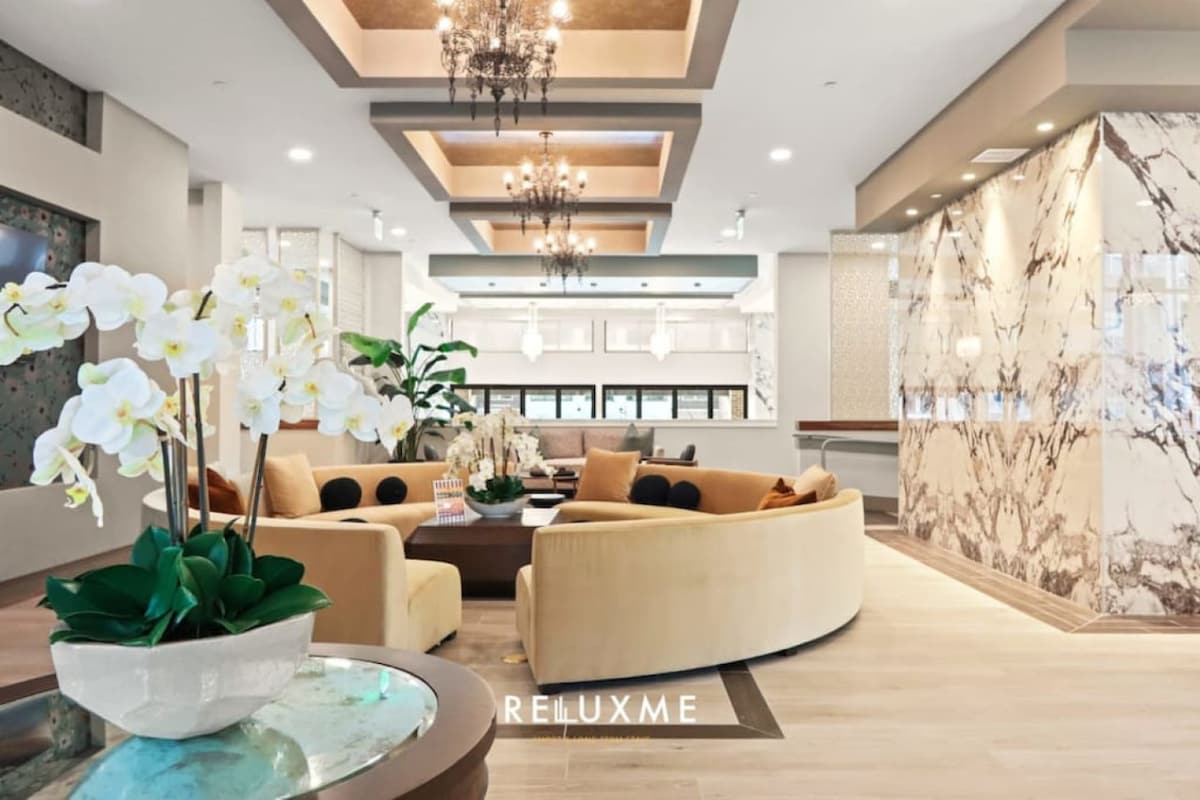 An elegant lobby area is shown, featuring a curved seating arrangement in soft beige upholstery. A round table is centered among decorative plants and white orchids. The space is adorned with chandelier lighting and a marble accent wall, creating a refined atmosphere.