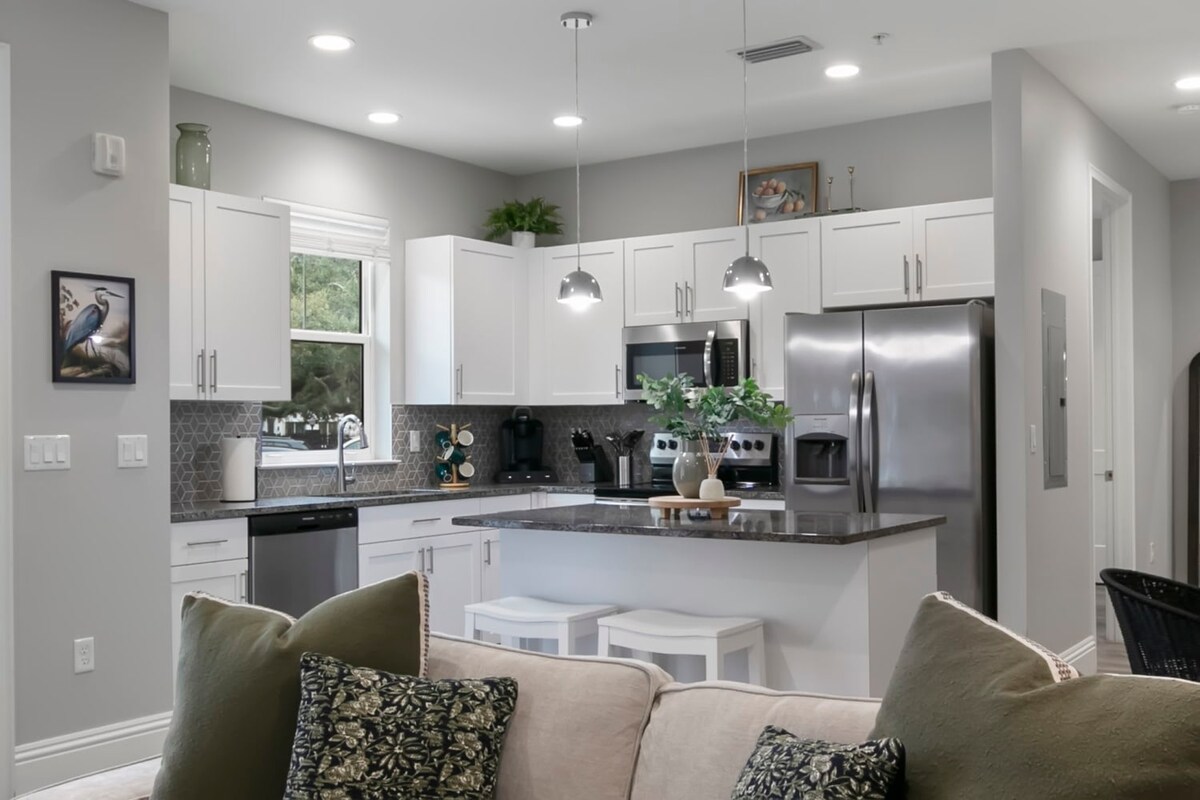 A modern kitchen features white cabinetry and stainless steel appliances. A dark countertop offers a contrast with the light decor, while stylish pendant lights hang overhead. Two bar stools provide seating at the kitchen island, creating an inviting space for meal preparation and casual dining.