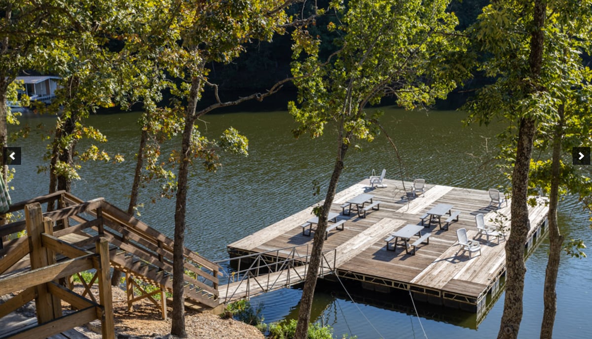A spacious community swim dock extends over the water, surrounded by leafy trees. Several seating areas with chairs offer views of the lake, inviting relaxation. A wooden staircase leads from the villa to the dock, enhancing accessibility to the outdoor space.