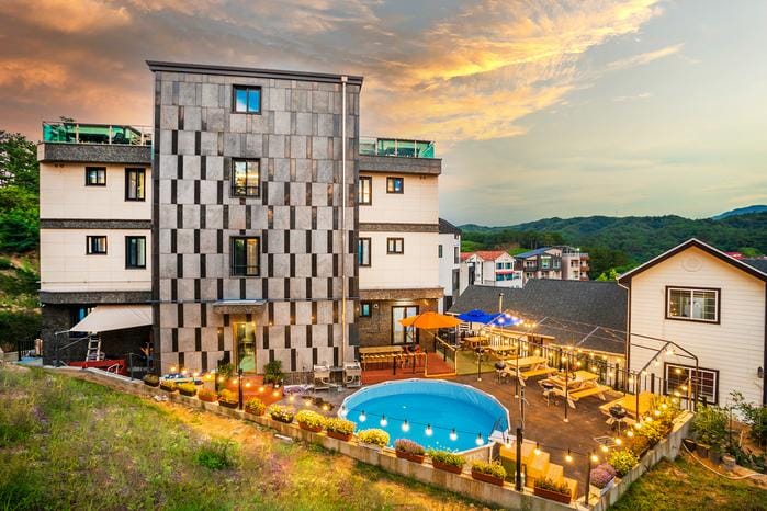 A modern building is positioned against an evening sky, featuring a circular outdoor pool surrounded by lounge seating and vibrant landscaping. Patio lights create a welcoming ambiance for outdoor gatherings. Multiple levels of the structure allow for a blend of indoor and outdoor experience.