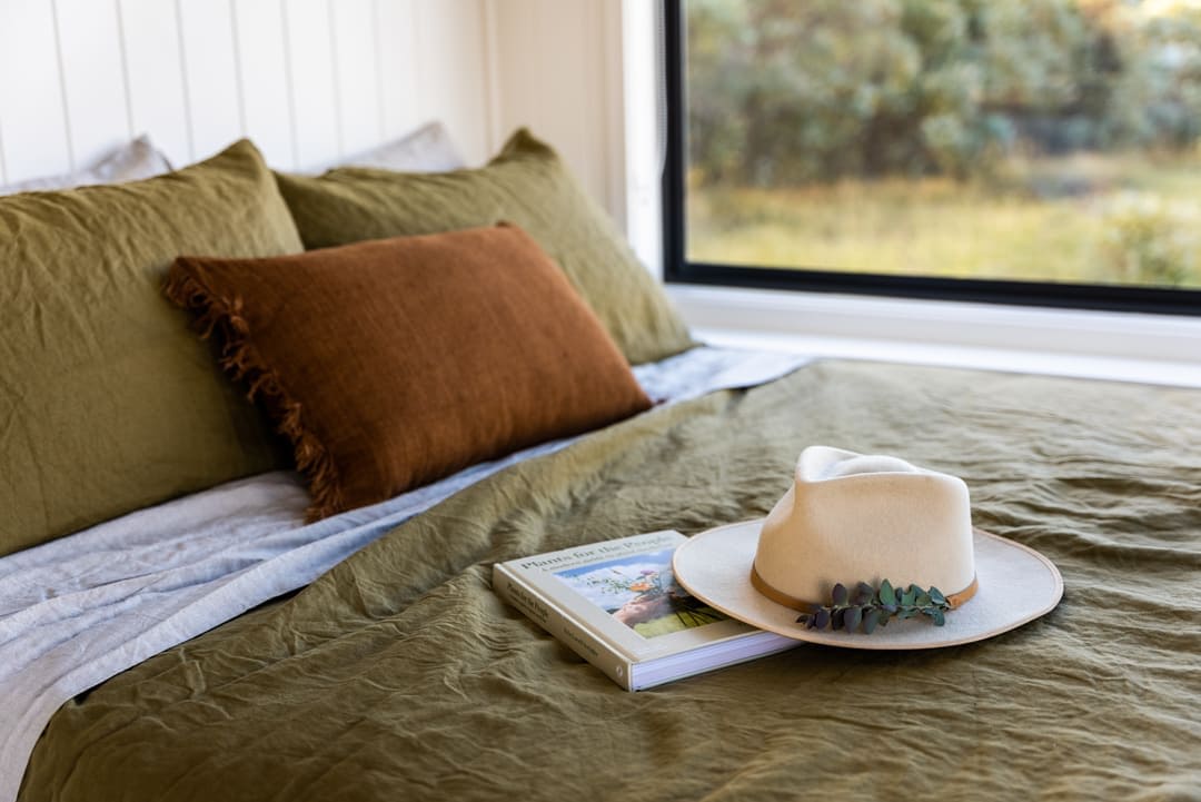 A spacious bed is dressed in earthy-toned linens, accented by a brown decorative pillow and a soft blanket. Nearby, a hat is placed atop a coffee table book, with a large window offering a glimpse of the natural landscape outside.