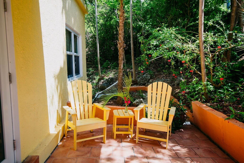 A small outdoor seating area features two yellow Adirondack chairs positioned beside a wooden table. Lush greenery and colorful flowers enhance the natural surroundings, creating a serene space for relaxation. Sunlight filters through the foliage, adding warmth to the area’s inviting atmosphere.