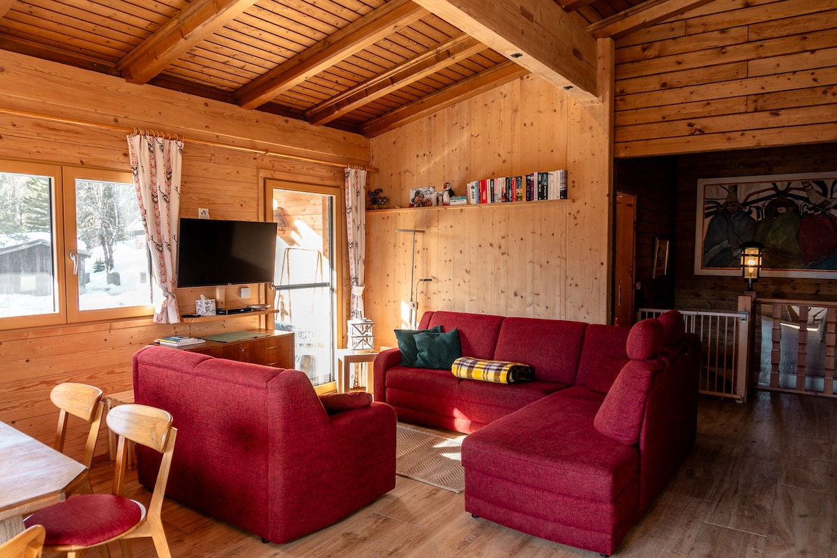 A spacious living room featuring a comfortable red sectional sofa and a cozy armchair. A wooden table with chairs is visible, alongside a television mounted on the wall. Natural light enters through large windows, illuminating the warm wooden interior.