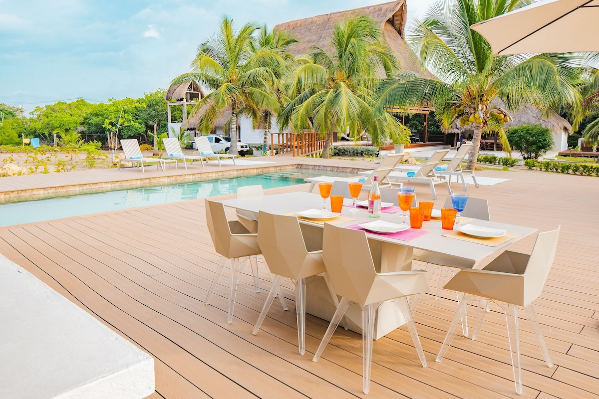 An outdoor dining area features a large table surrounded by modern chairs, set with colorful dishes and glasses. A private pool reflects the lush greenery, while lounge chairs and palm trees create a serene atmosphere. The area is bathed in natural light, enhancing the inviting environment.