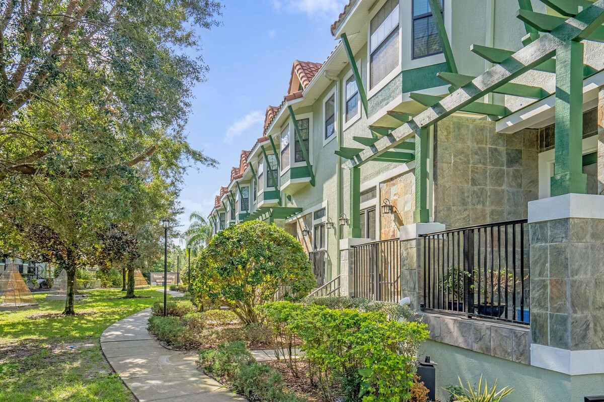 A row of split-level townhomes is set amidst neatly maintained gardens. Pathways wind through the landscape, leading to private porches with decorative railings, while large windows reflect the greenery surrounding the residences under a clear blue sky.