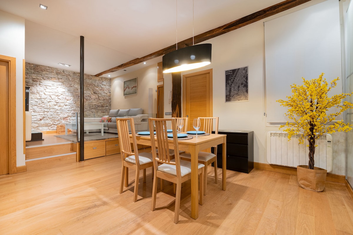 A dining area is highlighted by a wooden table surrounded by four chairs. A black pendant light suspends above, illuminating the space. A decorative yellow plant adds a colorful accent, and warm wood flooring complements the neutral tones of the adjacent living area.