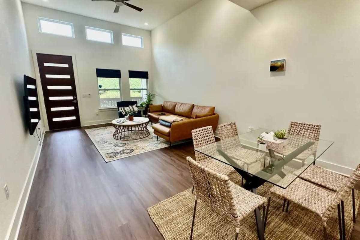 A bright living area features a comfortable brown leather sofa, accompanied by a glass coffee table atop a decorative rug. Six woven dining chairs surround a glass table, and large windows allow natural light to fill the room. A small plant adds a touch of greenery.