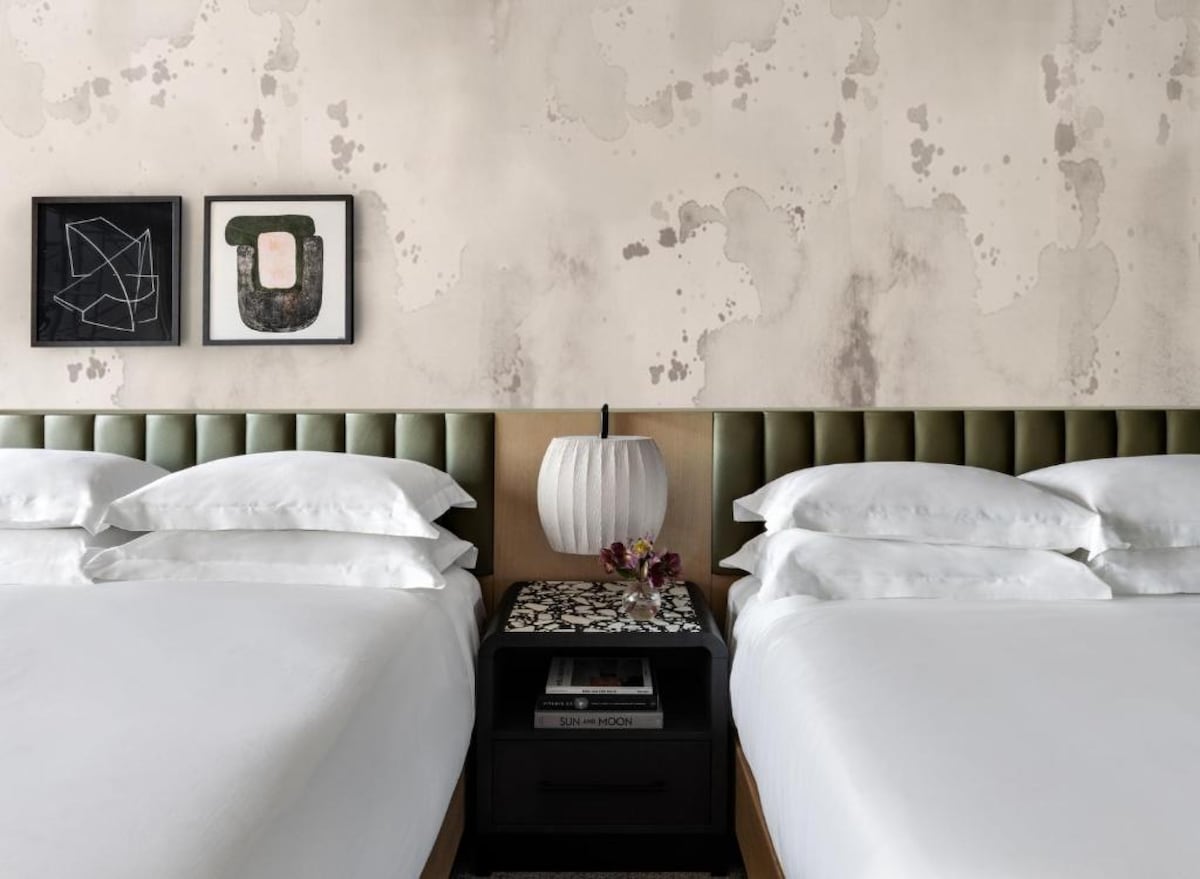 A room featuring two spacious queen beds with crisp white linens. A contemporary lamp is situated on a bedside table adorned with decorative items. Artwork hangs above the beds, complementing the softly textured walls.