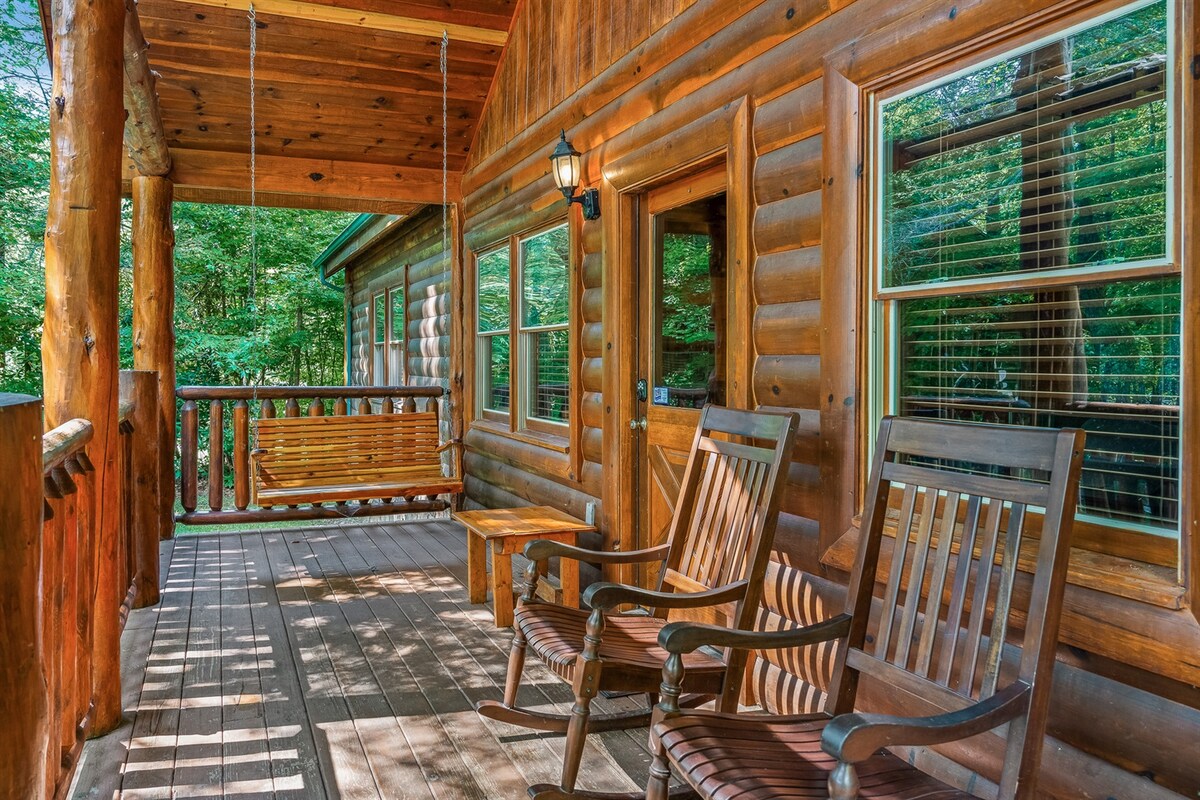 A spacious porch offers a serene retreat, featuring two wooden rocking chairs and a swing. Sunlight casts gentle shadows on the wooden deck, enhancing the rustic charm of the log cabin, while surrounding trees create a tranquil natural atmosphere.