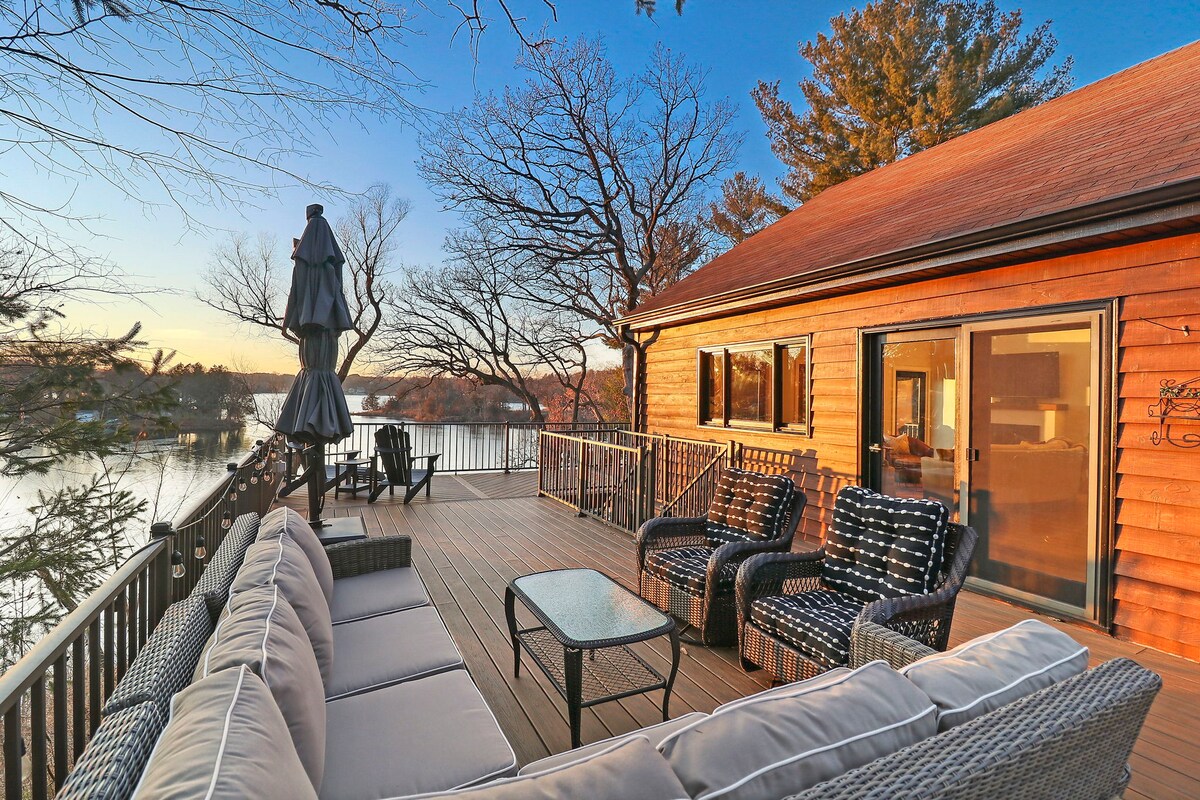 A spacious wraparound deck offers comfortable seating with black and white patterned cushions around a glass-topped table. The setting provides views of the lake and trees, enhancing the tranquil atmosphere during sunset.