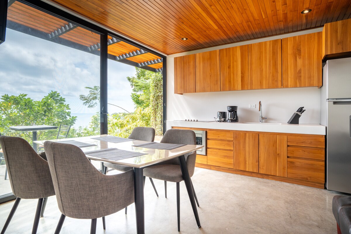 A modern kitchen area features wooden cabinetry and sleek appliances. A glass dining table is surrounded by four gray chairs, providing a comfortable space for meals. Large windows allow natural light to fill the room and showcase views of lush greenery.