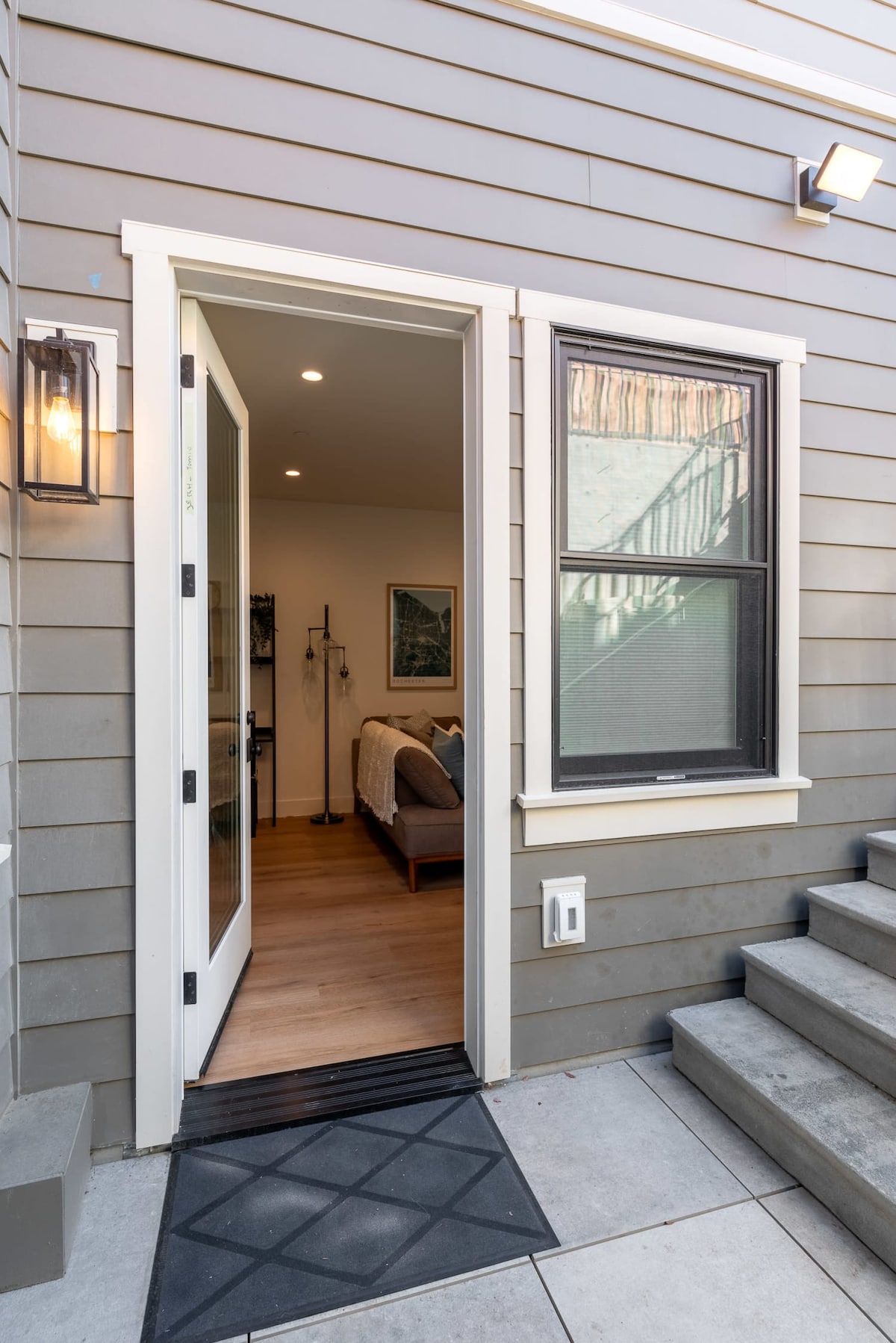 The entrance features a modern door framed by a sleek light fixture, opening to reveal a glimpse of the interior's warm design. A spacious staircase leads up from the entrance, accompanied by a large window that invites natural light into the space.