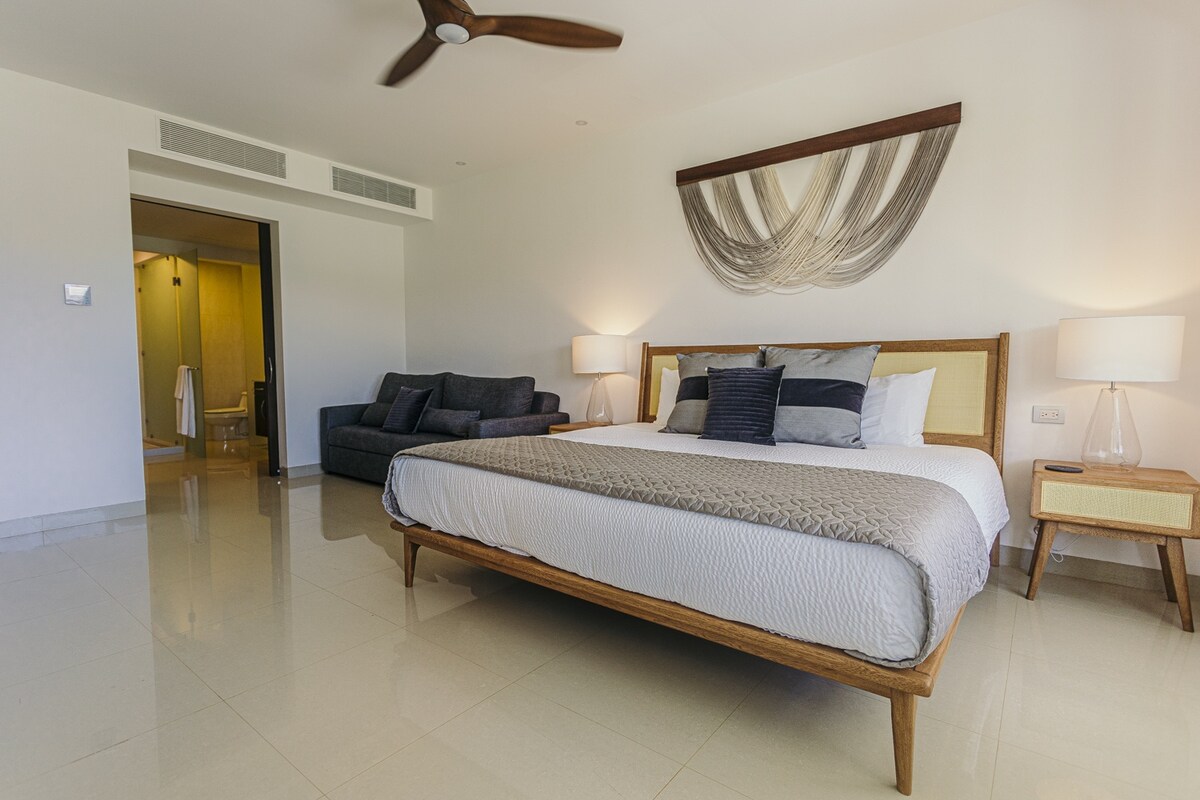 A spacious bedroom is presented with a large bed featuring a neutral-toned coverlet. A comfortable sofa is positioned in one corner, while soft lighting from bedside lamps enhances the serene environment. The room offers an open, uncluttered layout and a clean aesthetic.