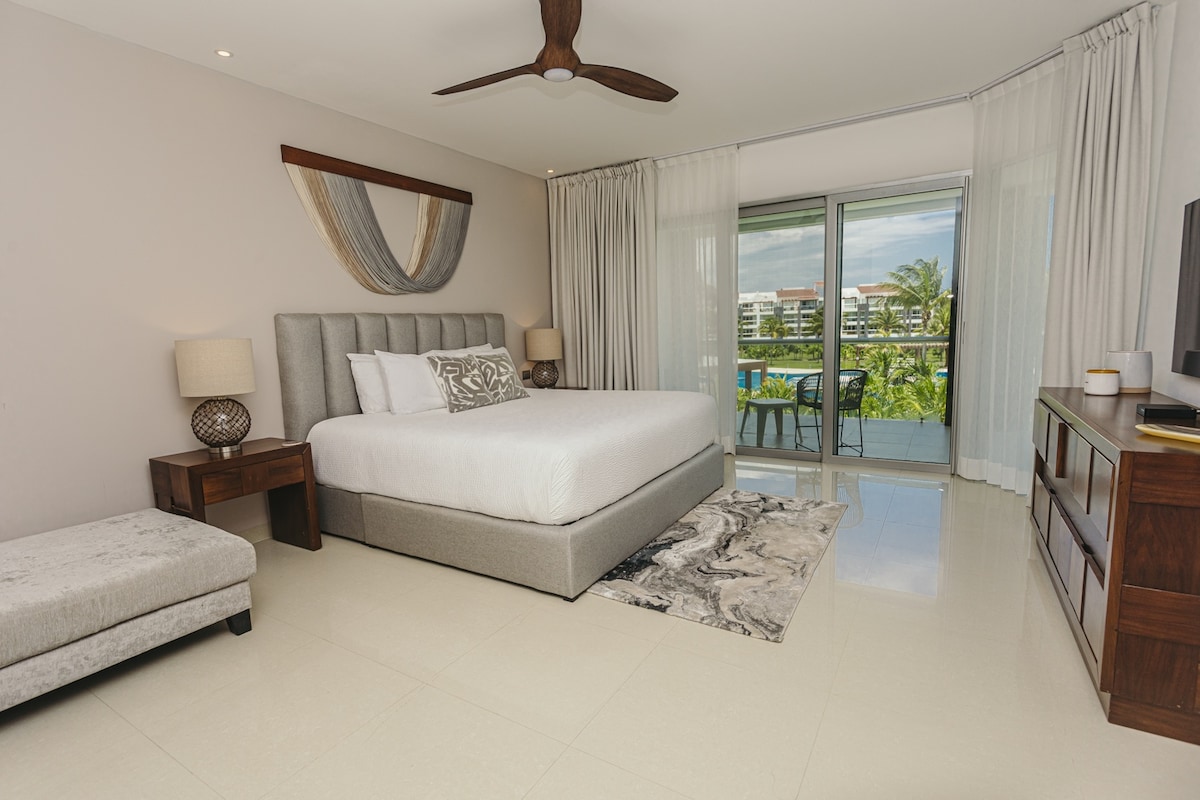 A well-furnished bedroom features a spacious bed with neutral bedding and a decorative wall hanging. Large glass doors lead to a balcony overlooking greenery, while soft natural light filters through sheer curtains. A sleek dresser and a small accent chair complete the inviting space.
